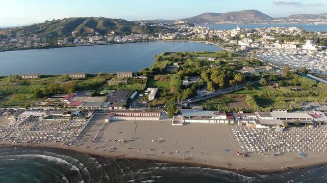 Miseno Beach Resort View Moving Over White Parasol Umbrellas and Across Sandy Coastline. Beautiful Mediterranean Marina Harbor Hidden Behind Green Peninsula Trees. Drone Shot