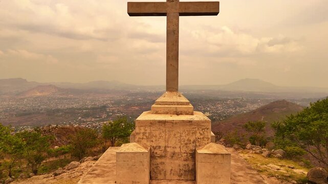 Drone rises from the base of the summit cross on Michiru Mountain, gradually revealing the full cross overlooking Blantyre from the Way of the Cross pilgrimage hike beneath a hazy sky.