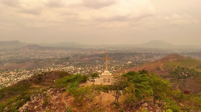 Drone pullback from the summit cross on the Way of the Cross trail atop Michiru Mountain, revealing panoramic views of Blantyre beneath a hazy, cloud-covered sky.