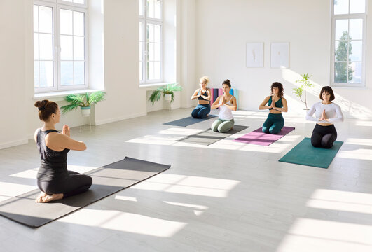Yoga class holds pose in bright studio. A diverse group kneels with hands at heart as the instructor guides calm breathing and alignment. Mindfulness, balance, and wellness through meditation.