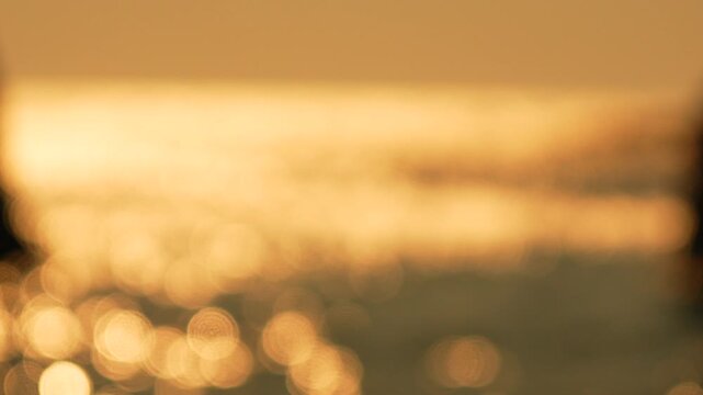 Golden light glimmers across moving ocean water at sunset creating a peaceful atmosphere near coastal rocks. Extreme soft focus static shot.