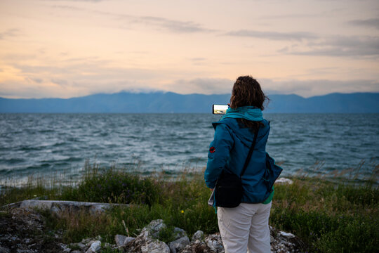 Female tourist taking landscape photograph of lake and mountains with smartphone at sunset