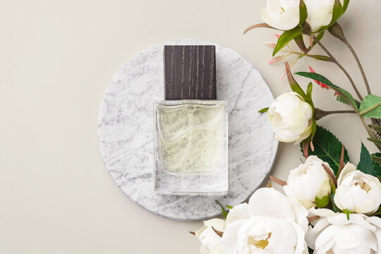 Perfume bottle on a round marble podium, decorated with decorative white roses and green leaves on beige background