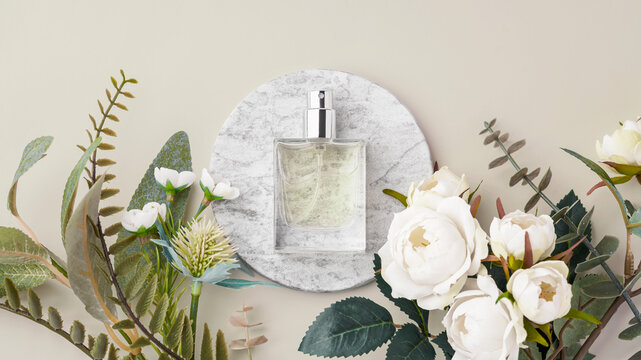 Perfume bottle on marble tray with white roses and green leaves decoration on beige background