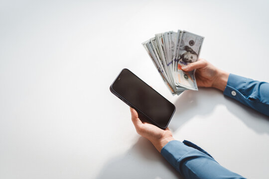 Person holding a smartphone in one hand and a stack of us dollar banknotes in the other, symbolizing mobile finance, online earning, and digital payment methods