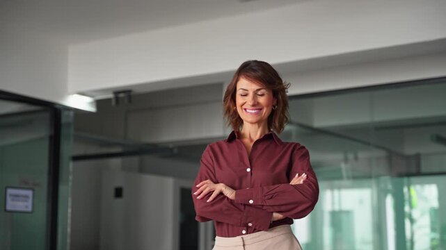 Portrait of confident businesswoman in 40s standing with arms crossed in office at copy space. Mature woman boss looks at camera with light smile. Trustworthy female business chief leads company.