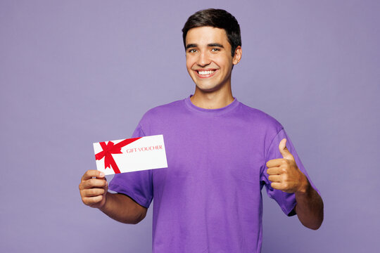Young man he wear violet t-shirt casual clothes hold gift certificate coupon voucher card for store show thumb up isolated on plain pastel light purple background studio portrait. Lifestyle concept.