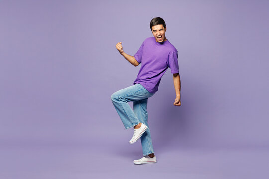 Full body young man wears violet t-shirt casual clothes doing winner gesture celebrate clenching fists say yes raise up leg isolated on plain pastel light purple background studio. Lifestyle concept.