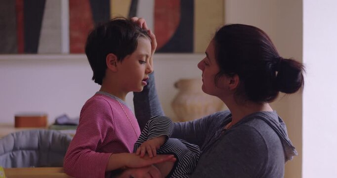 Caring mother caresses older child's hair while cradling toddler in her arms beside the family table at home, balancing sibling attention with tenderness and equality
