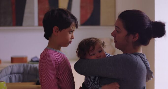 Attentive mother talks with older child while holding younger toddler in her arms at the dining table at home, sharing attention to foster security, fairness and sibling harmony