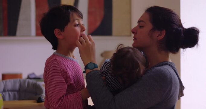 Child shows loose tooth as mother checks his mouth while holding younger sibling at home, marking childhood growth through attentive parenting and family care