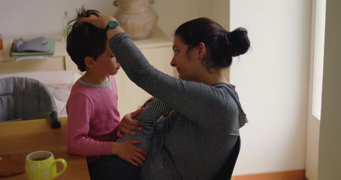 Mother caresses older son's hair while breastfeeding toddler at the breakfast table at home, balancing sibling attention with tenderness, reassurance and inclusive family care