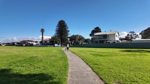A person walking a dog along pathway in a lush green reserve in Seaholme, Melbourne, Australia. A liveable suburban environment, relaxed local lifestyle, outdoor recreation, and peaceful neighborhood