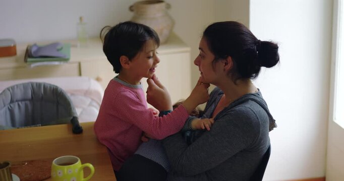 Mother and young son hold each other's chins and try not to smile while toddler rests in her arms at home, sharing authentic laughter, playful bonding and family joy