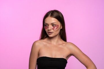 Fototapeta premium Young woman applying cosmetic eye patches for under eye skin care, looking away on pink studio background