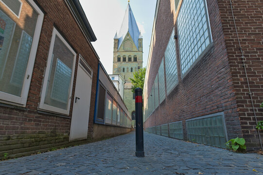 A narrow cobblestone alley stretches between brick buildings toward a distant church tower with a blue spire. A black and red bollard stands in the alley - Soest, Westphalia, Germany