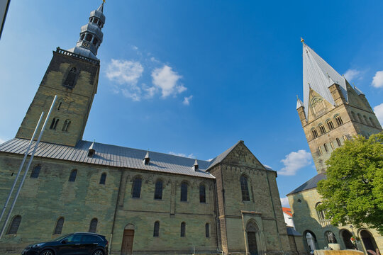 Two striking historic church towers stand against a bright blue sky. A tall, dark stone tower is on the left, with a grander cathedral tower and spire on the right - Soest, Westphalia, Germany