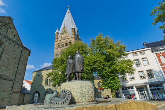 A historic European town square features a sculpture of two people, framed by trees, in front of a prominent church with a tall, pointed spire - Soest, Westphalia, Germany