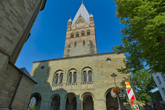 An imposing historical stone building with a tall tower and pointed spire stands against a blue sky, flanked by green trees - Soest, Westphalia, Germany