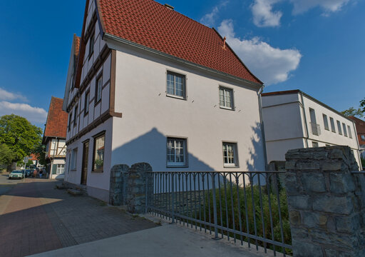 A two-story white building with a red roof stands next to a half-timbered house under a blue sky. A low metal fence is in the foreground - Soest, Westphalia, Germany