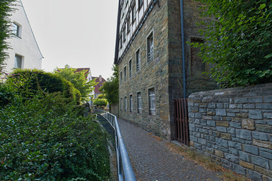 A narrow, sunlit pathway leads past an old stone building with small windows. Green foliage and a metal railing line the path in this European street scene - Soest, Westphalia, Germany