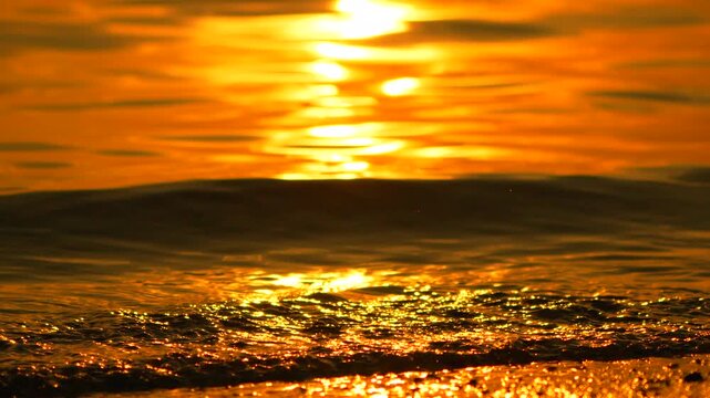 Vibrant orange sunset glow illuminates rhythmic sea tide as water gently washes across the sandy coastline. Static macro shot.