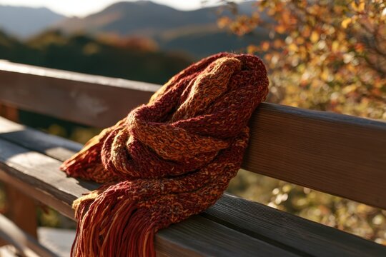 Warm multicolored knitted scarf resting on a wooden bench surrounded by the peaceful atmosphere of an autumn mountain setting.
