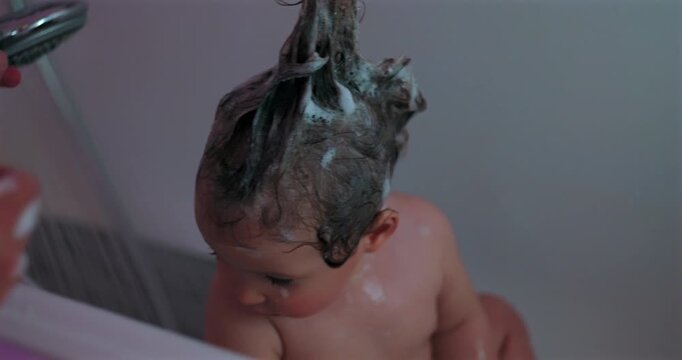 Parent shampoos toddler&rsquo;s hair in bathroom tub, pulling soapy strands upward into a funny hairstyle during playful bathtime, hygiene care and family bonding