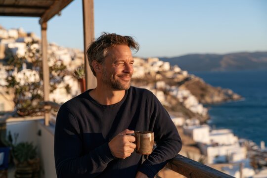 Smiling mature man enjoying a morning coffee on a balcony with panoramic views of a charming seaside town and the Mediterranean coastline.
