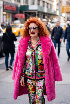 Fashionable cheerful senior woman with bright red curly hair confidently walks down a bustling city street wearing a vibrant pink fluffy coat, colorful floral suit and sunglasses.