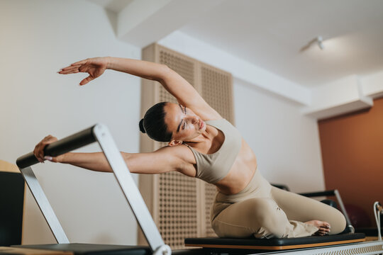 A woman performs a pilates reformer workout, stretching on the machine in a bright studio. She leans sideways while holding the reformer bar for a controlled flexibility exercise.