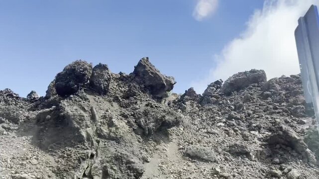 Dynamic side window view of driving car passing volcanic rocky hillside in Tenerife. Deserted mountain slope with dry wild shrubs, metal guardrail bar on foreground under cloudy sky. Summer roadtrip.