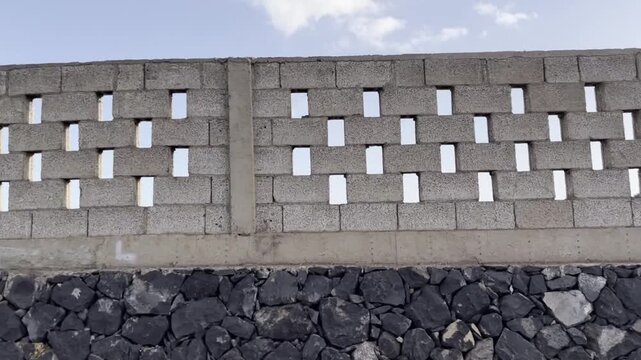 Dynamic side window view of driving passenger passing long grey cinder block brick fence with decorative gaps in Tenerife. Geometric concrete stone wall texture under bright sunny blue sky with clouds