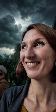 Portrait einer dunkelhaarigen Frau, sie schaut lachend nach oben, dunkle Wolken am Himmel, ein Gewitter zieht herauf und es regnet