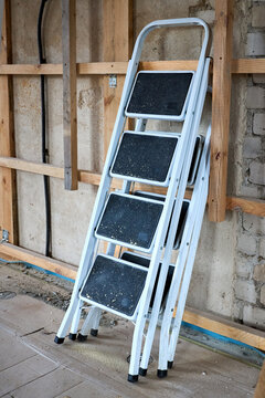 White metal 4-step folding ladder stands near wooden framing on dusty concrete floor of house under renovation. Used portable equipment with nonslip steps for construction and home repair.
