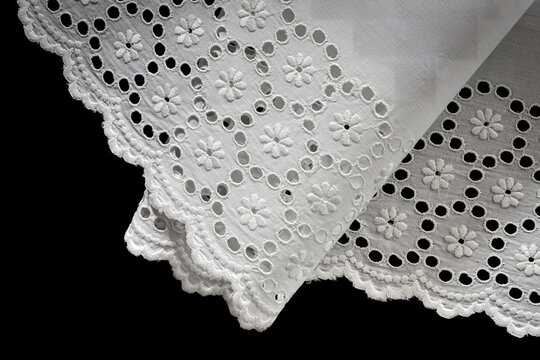 Draped and folded corner of white cotton broderie anglaise lace trim, revealing the eyelet floral pattern and scalloped hem from two angles, isolated on black background.