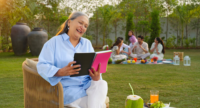 Indian old aged relax woman mom sitting on chair hold book read story novel outdoor house place happy male female man group talk speak spend quality time enjoy new trip day open in air garden lawn