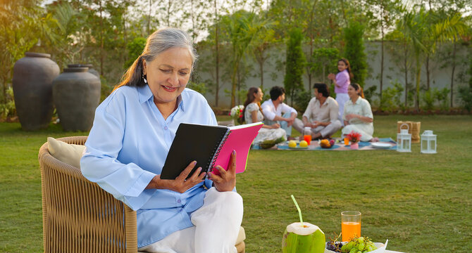 Indian old aged relax woman mom sitting on chair hold book read story novel outdoor house place happy male female man group talk speak spend quality time enjoy new trip day open in air garden lawn