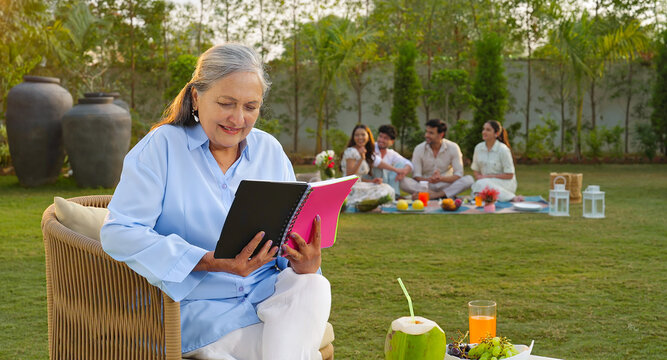 Indian old aged relax woman mom sitting on chair hold book read story novel outdoor house place happy male female man group talk speak spend quality time enjoy new trip day open in air garden lawn