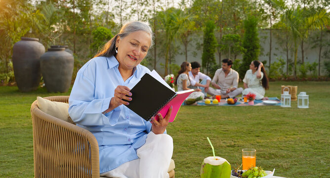 Indian old aged relax woman mom sitting on chair hold book read story novel outdoor house place happy male female man group talk speak spend quality time enjoy new trip day open in air garden lawn