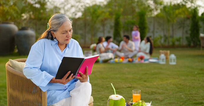 Indian old aged relax woman mom sitting on chair hold book read story novel outdoor house place happy male female man group talk speak spend quality time enjoy new trip day open in air garden lawn
