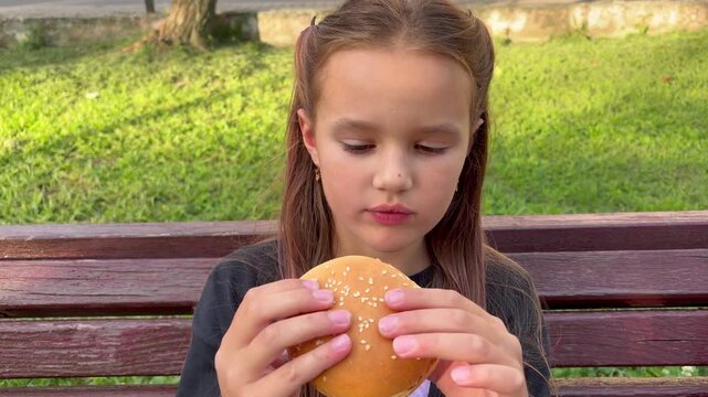 A 7-8 year old girl eats a cheeseburger in a city park.