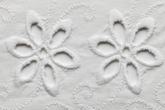 Macro shot of two six-petal cutwork daisy flowers embroidered on white cotton sangallo fabric, shown against a soft light background.