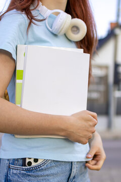 Close-up of a teenage girl carrying a white notebook under her arm with headphones around her neck while walking outdoors. Concept of education, student lifestyle and back to school.
