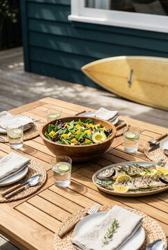 Beautiful summer outdoor dining table set with healthy fresh salad, grilled fish and cold drinks on a wooden deck. A yellow surfboard leans against a blue beach house in the bright sunlight.