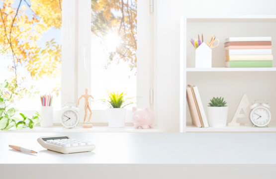 Modern workspace white table top with stationery, calculator and book shelf on blurred window sill background