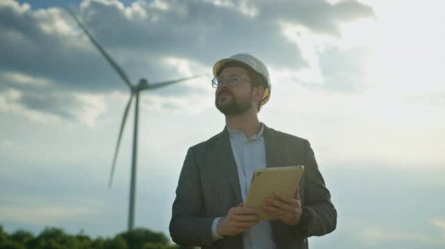Man in hard hat and suit holds tablet computer in front of wind turbines on cloudy day. Renewable energy, clean electricity, wind power inspection, eco-friendly technology, sustainable industry,