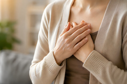 Young woman holding hands on chest feeling heart pain or stress sitting in bright home interior view.
