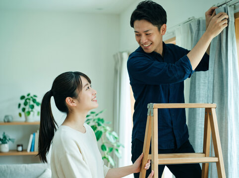 カーテン設置中に脚立を支えながらパートナーと視線を交わす笑顔の女性