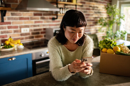 Woman using smartphone in kitchen with fresh grocery delivery